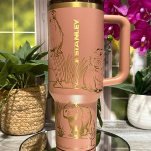 Puede incluir: Vaso térmico Stanley de color rosa con una ilustración dorada de carpinchos y juncos. El vaso tiene un asa y una tapa transparente. El logotipo de Stanley está impreso verticalmente en dorado.