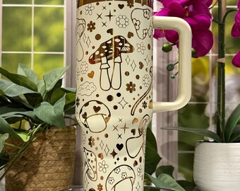 May include: A cream-coloured insulated tumbler with a gold mushroom pattern and a cream-coloured handle. The tumbler has a clear lid and is decorated with various mushroom designs, flowers, and stars. The tumbler is approximately 25 cm tall.
