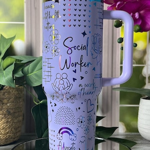 May include: A lilac tumbler with a handle, decorated with the words "Social Worker" and "Advocate." The design includes floral and heart motifs, and the phrase "because mental health matters." It has a silver rim and a lilac lid.