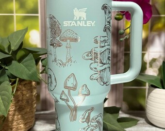 May include: A light teal Stanley tumbler with a white lid and handle. The tumbler features a gold illustration of mushrooms and the Stanley logo. A white straw is inserted into the lid. The tumbler is sitting on a mirrored surface.