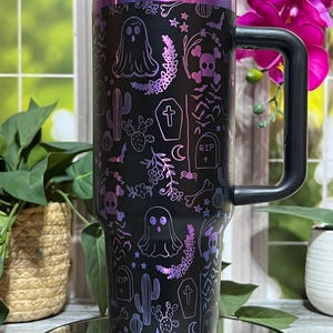 Könnte beinhalten: Schwarzer Reisebecher mit Henkel, mit einem Halloween-Motiv. Das Design umfasst Geister, Totenköpfe, Kakteen und Grabsteine mit einem lila und rosa Farbverlauf. Der Becher hat einen schwarzen Deckel und Henkel.