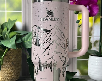 May include: A pink Stanley tumbler with a handle and straw. The tumbler features a mountain and Bigfoot design in silver. The Stanley logo is visible. The tumbler is approximately 25 cm tall.