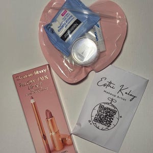 Puede incluir: Una colección de productos de belleza, que incluye un kit de labios, toallitas desmaquillantes y un pequeño recipiente de polvo. El kit de labios está en una caja rosa con un delineador de labios y un lápiz labial. Las toallitas desmaquillantes son azules y blancas. También se incluye un pequeño paquete blanco con un código QR.
