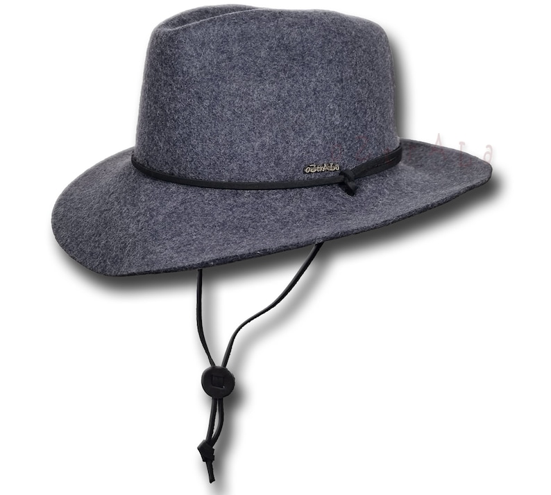 May include: A gray felt fedora hat with a black leather band and a black cord chin strap. The hat has a wide brim and a small, round crown. The brand name "Outback" is embossed on the leather band.