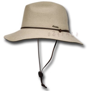 May include: A beige felt fedora hat with a brown leather band and a brown leather chin strap. The hat has a wide brim and a small, round crown.