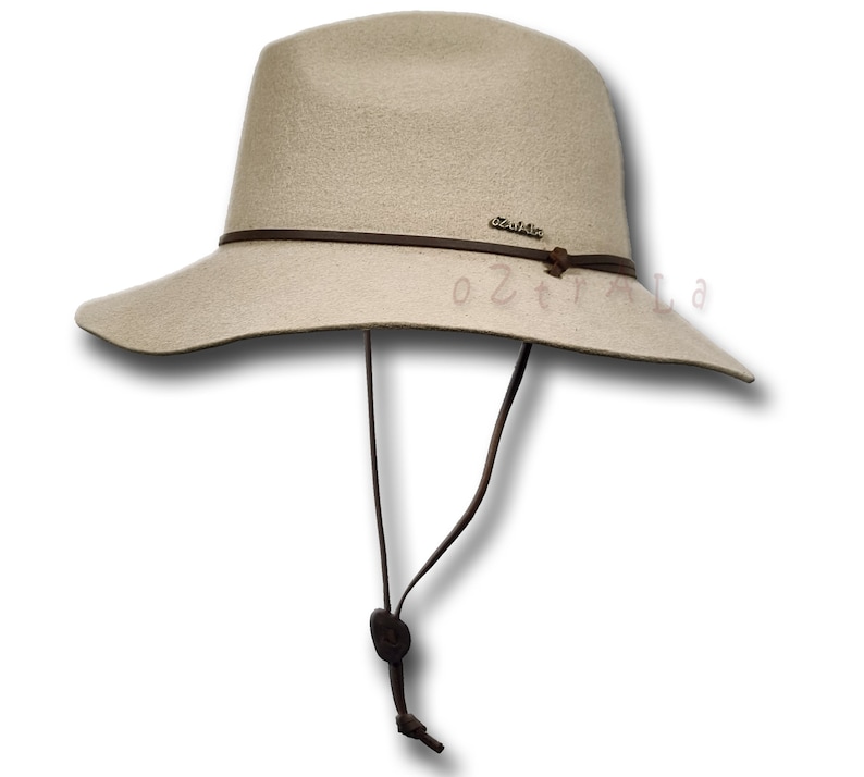 May include: A beige felt fedora hat with a brown leather band and a brown leather chin strap. The hat has a wide brim and a small, round crown.