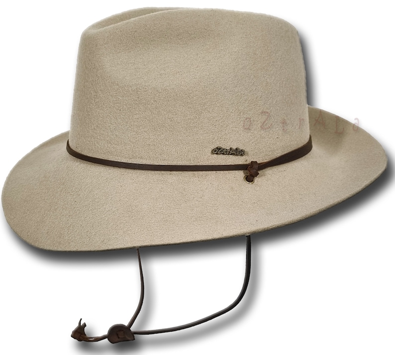 May include: A beige felt fedora hat with a brown leather band and a brown leather chin strap. The hat has a small metal tag that says "Outback" on it.