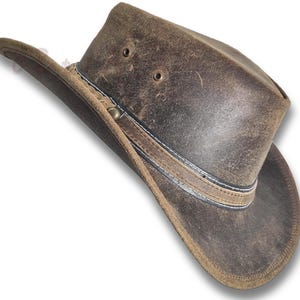 Puede incluir: Sombrero de vaquero de cuero marr&oacute;n desgastado con ala ancha. El sombrero presenta una banda decorativa con costuras blancas y detalles met&aacute;licos. Tiene dos orificios de ventilaci&oacute;n en la corona.