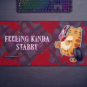 May include: A red and white mousepad with a cartoon cat holding a knife. The cat is orange and white with blue eyes. The text "Feeling Kinda Stabby" is written in pink letters on the mousepad.
