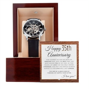 May include: A silver-toned skeleton watch with a black leather band is displayed in a wooden gift box. The watch has Roman numeral hour markers and visible internal gears. A card with the text "Happy 35th Anniversary" is included.