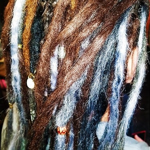 May include: Dreadlocks with brown, blue, and white synthetic hair extensions. The dreadlocks are decorated with gold metal beads and rings.