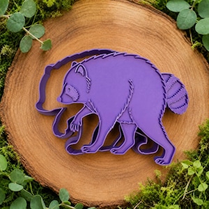 Puede incluir: Un cortador de galletas morado con forma de mapache que presenta un contorno detallado del animal, incluyendo su característico rostro enmascarado, orejas puntiagudas y cola rayada, diseñado para cortar masa con la forma de un mapache.