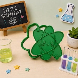 May include: A green atom-shaped cookie cutter featuring a smiling face with circular eyes and a small mouth. The design includes orbital lines crossing the center. The cutter is positioned among science-themed items including a small chalkboard sign.