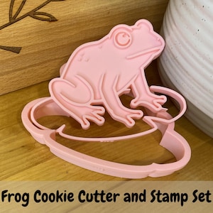 May include: A light pink plastic cookie cutter and stamp set featuring a frog sitting on a lily pad. The design includes a detailed outline of the frog with defined eyes, legs, and feet, along with the rounded shape of the lily pad base.
