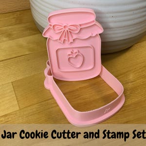 May include: A light pink plastic cookie cutter and stamp set shaped like a mason jar. The stamp features a lid with a bow detail and a central rectangular label containing a small strawberry icon. The cutter has a deep rim for cutting dough.