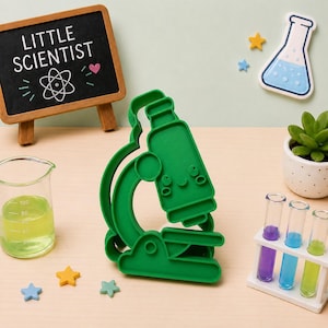 May include: A green plastic microscope-shaped cookie cutter with a smiling face. Nearby are a small chalkboard with the text LITTLE SCIENTIST, a beaker with yellow liquid, a test tube rack with three colored liquids, and small star-shaped decorations.