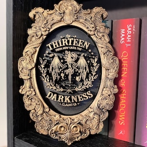 K&ouml;nnte beinhalten: Verziertes, goldfarbenes Wanddekor mit dem Text "the THIRTEEN FROM NOW UNTIL THE DARKNESS CLAIMS US+" auf schwarzem Hintergrund. Der ovale Rahmen steht auf einem schwarzen Regal neben B&uuml;chern.