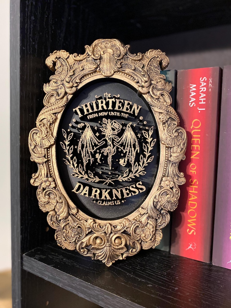 K&ouml;nnte beinhalten: Verziertes, goldfarbenes Wanddekor mit dem Text "the THIRTEEN FROM NOW UNTIL THE DARKNESS CLAIMS US+" auf schwarzem Hintergrund. Der ovale Rahmen steht auf einem schwarzen Regal neben B&uuml;chern.