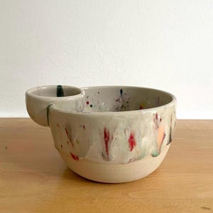 Könnte beinhalten: Eine Keramikschale mit einem kleineren, seitlich angebrachten Fach, die eine cremefarbene Basis mit bunten Farbspritzern in Rot, Blau und Orange aufweist. Die Außenseite hat eine glänzende Glasur, während der untere Teil unglasiert bleibt.