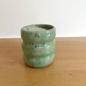 Könnte beinhalten: Ein hellgrüner Keramik-Zahnbürstenhalter mit gesprenkelter Oberfläche. Der Halter hat ein einzigartiges, gestapeltes Design mit einer geschwungenen Oberseite und einem kleinen Loch. Das handgefertigte Keramikstück ist ein dekoratives Badezimmerzubehör.