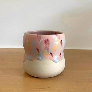 Könnte beinhalten: Ein Keramikbecher mit abgerundeter Form, einer glatten, matten cremefarbenen Basis und einer glänzenden, tropfenden Glasur in Pastellrosa, Hellblau und tiefem Magenta, die vom Rand bis zur Mitte des Gefäßes herabfließt.