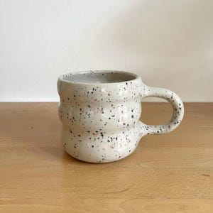 Könnte beinhalten: Eine gesprenkelte Keramiktasse mit rundem Körper und gebogenem Henkel. Die Tasse ist cremefarben mit schwarzen und rotbraunen Sprenkeln. Die Tasse steht auf einer Holzoberfläche.