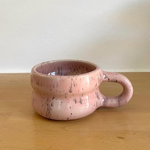 Pode incluir: Uma caneca de cerâmica curta e larga com um corpo arredondado e bojudo e uma pega grossa e curva. A caneca apresenta um acabamento de esmalte rosa claro brilhante com manchas cinzentas escuras ou pretas distribuídas por toda a superfície.