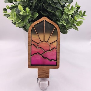 May include: A wooden night light shaped like a window with a sunset scene inside. The scene features a yellow sun, pink and orange mountains, and a dark blue sky.