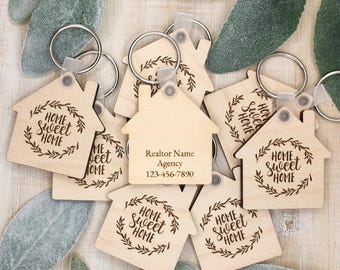 May include: A collection of wooden house-shaped keychains. The keychains are engraved with the phrase "Home Sweet Home" surrounded by a wreath design. One keychain has "Realtor Name, Agency, 123-456-7890" engraved on it. Each keychain has a silver keyring.