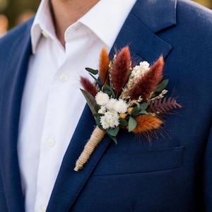 Könnte beinhalten: Ein marineblauer Blazer mit einem weißen Hemd und einem Knopfloch. Das Knopfloch zeigt getrocknete Blumen in Braun-, Orange- und Weißtönen, umwickelt mit Bindfaden. Der Blazer hat eine klassische Passform und ein fallendes Revers.