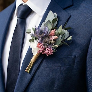 Op de afbeelding: Een marineblauw pak met een wit overhemd en een donkerblauwe stropdas. Een corsage van gedroogde bloemen in tinten paars, roze en groen is op de colbertjas gespeld. De corsage is omwikkeld met touw.