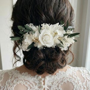 May include: A white floral hair comb featuring a large rose, small white flowers, and green leaves. The hair accessory is set against dark brown hair and a white lace dress, perfect for a wedding or special occasion.