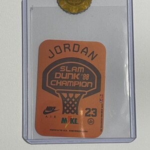 Puede incluir: Tarjeta de baloncesto coleccionable con Michael Jordan. La tarjeta tiene un fondo naranja con las palabras "JORDAN" y "SLAM DUNK '88 CHAMPION" en negro. El logotipo de Nike Air y el n&uacute;mero 23 tambi&eacute;n son visibles.