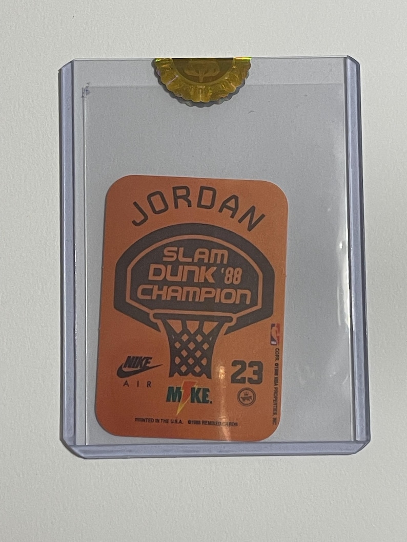 Puede incluir: Tarjeta de baloncesto coleccionable con Michael Jordan. La tarjeta tiene un fondo naranja con las palabras "JORDAN" y "SLAM DUNK '88 CHAMPION" en negro. El logotipo de Nike Air y el n&uacute;mero 23 tambi&eacute;n son visibles.