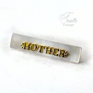 Op de afbeelding: Witte rechthoekige haarspeld met het woord "MOTHER" in gouden letters.