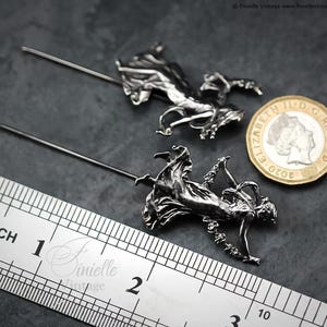 May include: Two silver metal hat pins, each with a detailed figure of a woman in flowing attire. A ruler shows the pins are approximately 5 cm long. A coin is next to the pins for size comparison.
