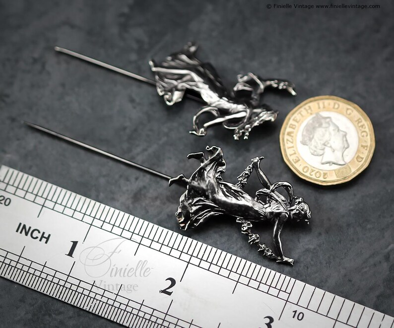 May include: Two silver metal hat pins, each with a detailed figure of a woman in flowing attire. A ruler shows the pins are approximately 5 cm long. A coin is next to the pins for size comparison.