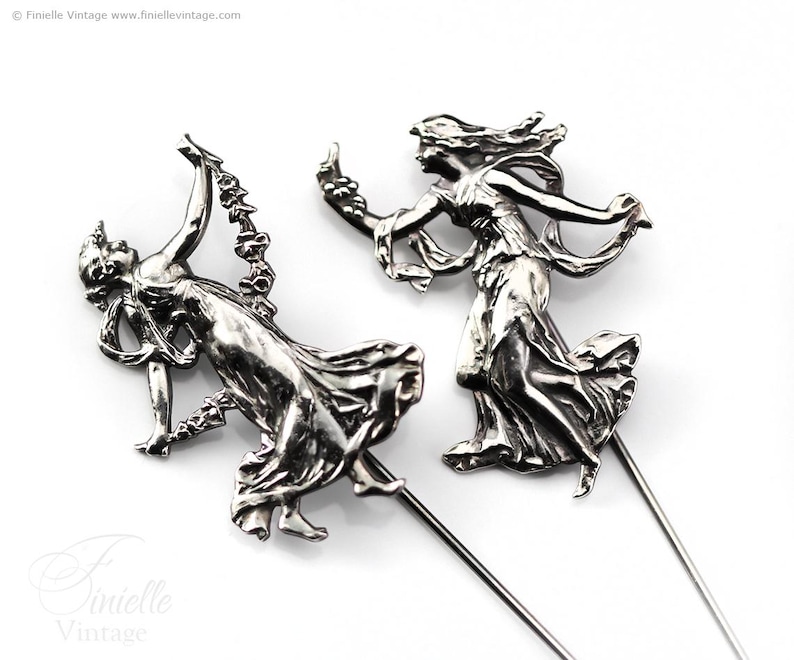 May include: Two silver-toned metal hat pins depicting classical female figures in flowing gowns. The figures are in dynamic poses, with one holding a floral garland. The pins have long, slender shafts.