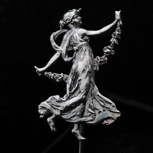 May include: A silver sculpture of a woman in a flowing dress, holding a garland. The figure is in a dynamic pose, with one leg raised and arms outstretched. The sculpture is mounted on a thin metal rod against a black backdrop.