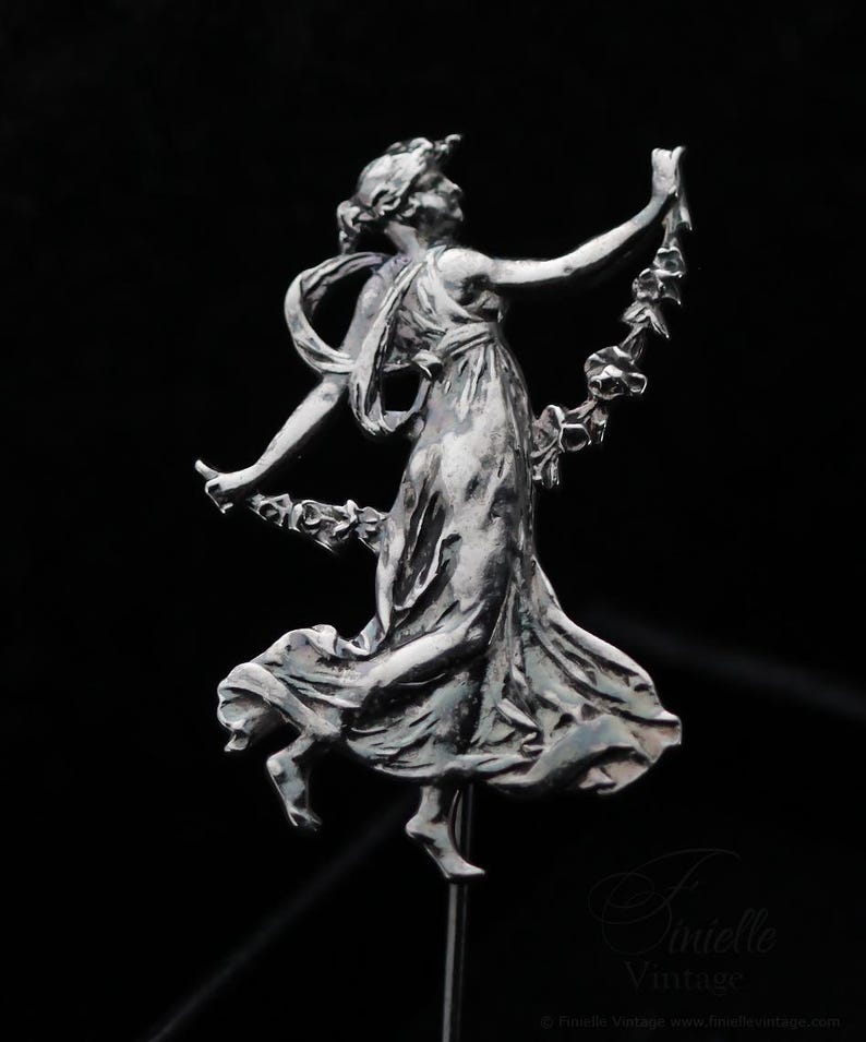 May include: A silver sculpture of a woman in a flowing dress, holding a garland. The figure is in a dynamic pose, with one leg raised and arms outstretched. The sculpture is mounted on a thin metal rod against a black backdrop.