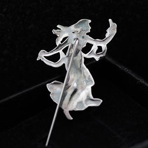 May include: A silver-coloured vintage brooch depicting a dancing woman. The pin features intricate detailing with flowing lines and curves, showing a figure with outstretched arms and a flowing dress. The brooch is set against a dark background.