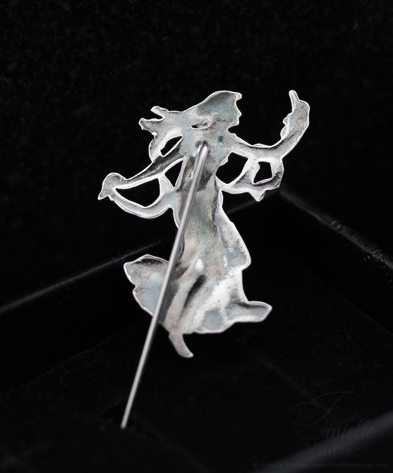 May include: A silver-coloured vintage brooch depicting a dancing woman. The pin features intricate detailing with flowing lines and curves, showing a figure with outstretched arms and a flowing dress. The brooch is set against a dark background.