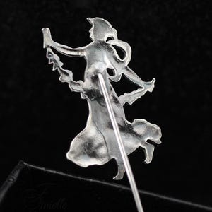 May include: A silver-coloured vintage brooch pin depicting a stylised female figure in a flowing dress, holding an object. The pin has a long, straight needle for attachment. The design is intricate and detailed.