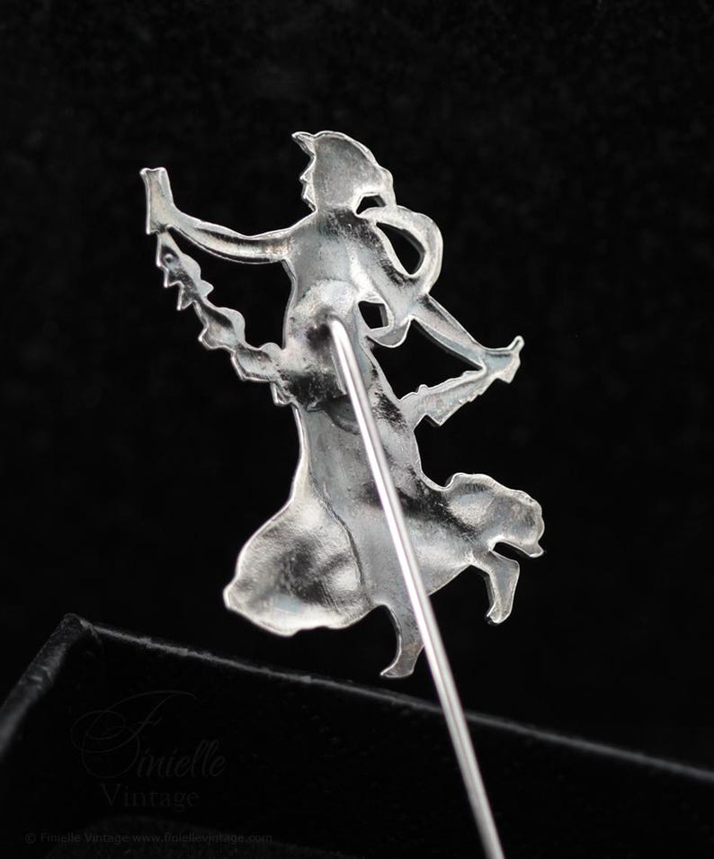 May include: A silver-coloured vintage brooch pin depicting a stylised female figure in a flowing dress, holding an object. The pin has a long, straight needle for attachment. The design is intricate and detailed.