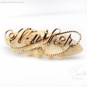Op de afbeelding: Een goudkleurige draadsculptuur met het woord "Mother" in cursief schrift, rustend op een lichtgekleurde, getextureerde basis. Het woord is vervaardigd met elegante krullen en een gedraaide draadrand, waardoor een delicaat en artistiek stuk ontstaat.
