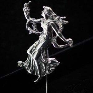 May include: A silver metal sculpture of a woman in a flowing dress, arms extended, holding a curved item. The figure is mounted on a thin metal rod against a black backdrop. The sculpture has detailed features and a vintage style.