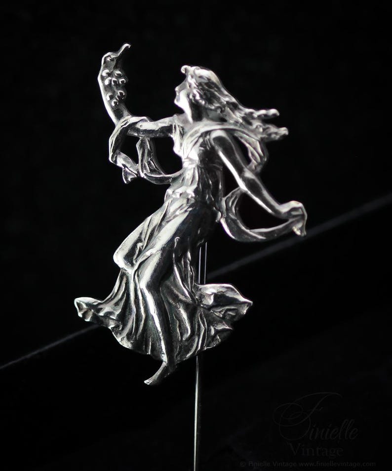 May include: A silver metal sculpture of a woman in a flowing dress, arms extended, holding a curved item. The figure is mounted on a thin metal rod against a black backdrop. The sculpture has detailed features and a vintage style.