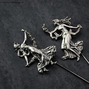 May include: Two silver hat pins depicting classical female figures in flowing gowns. The figures are in dynamic poses, with one holding a garland and the other reaching upwards. The pins have long, slender shafts.