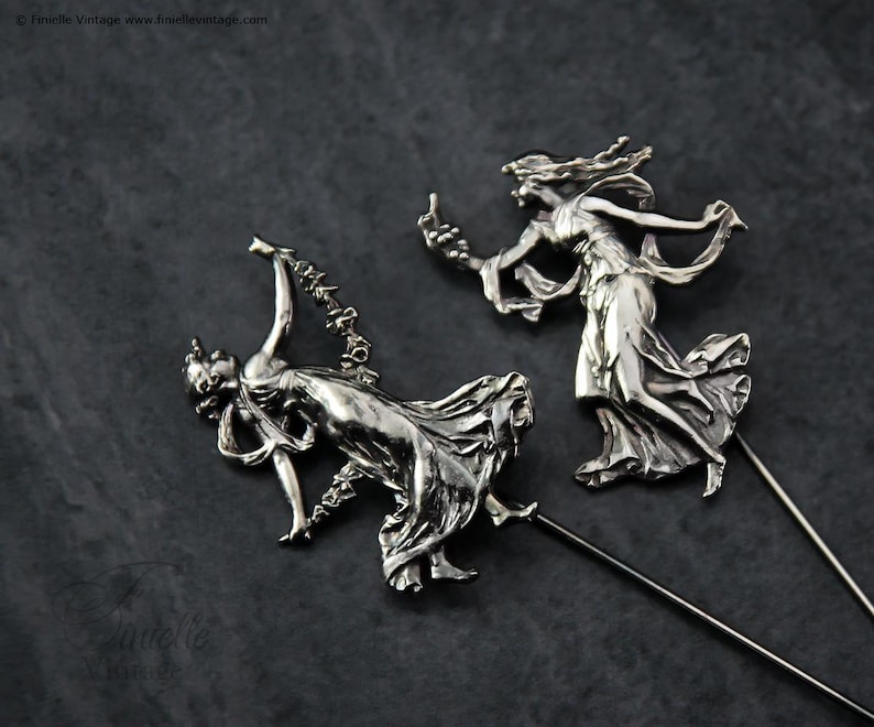 May include: Two silver hat pins depicting classical female figures in flowing gowns. The figures are in dynamic poses, with one holding a garland and the other reaching upwards. The pins have long, slender shafts.