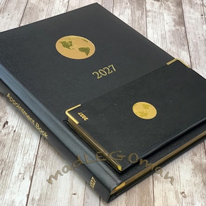 May include: A large black appointment book and a smaller matching black planner, both featuring gold foil globe icons and the year 2027 printed in gold. The larger book has the words Appointment Book printed in gold on the side spine.